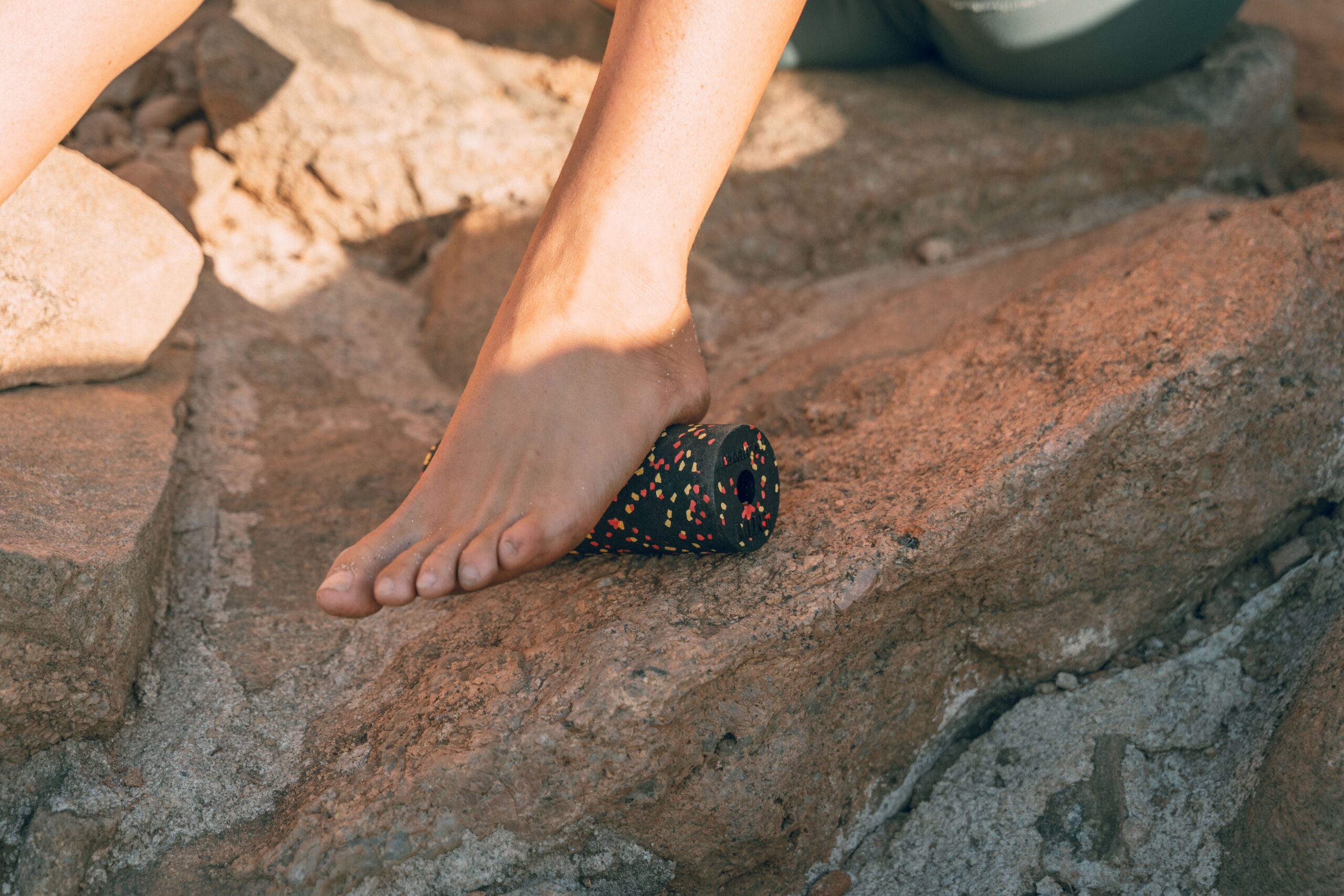 Close-up of a foot using a massage roll on rocky terrain outdoors in warm light.